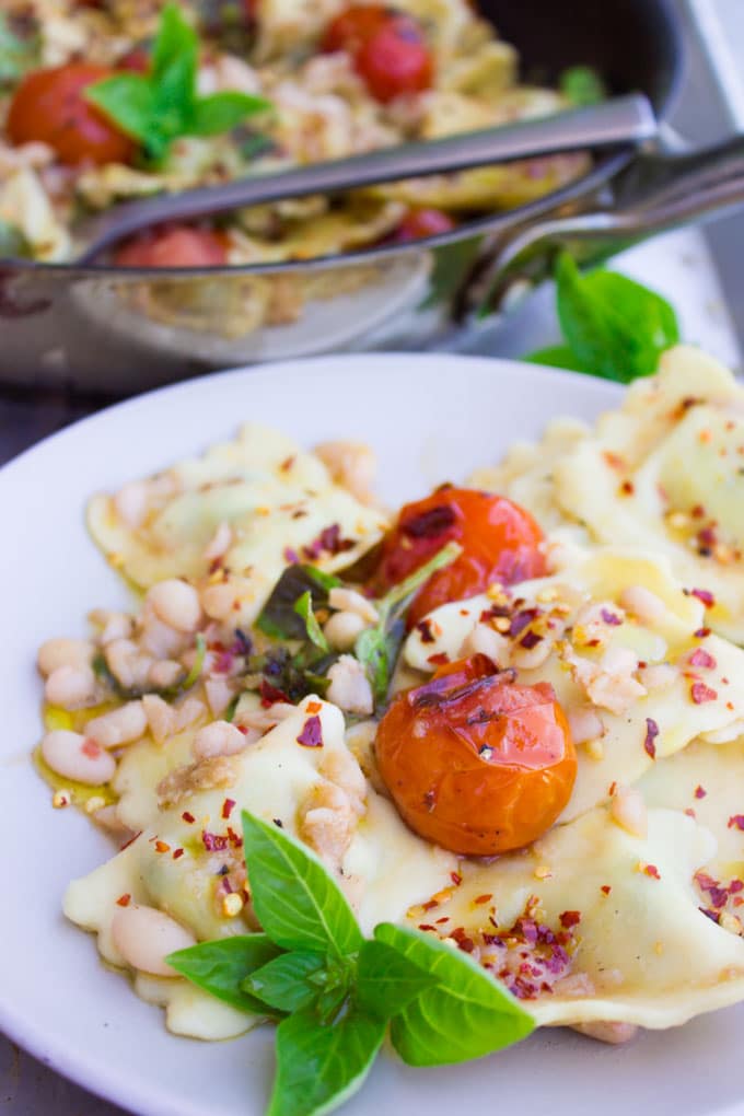 close-up of a serving of 15 Minute One Pan Ravioli with navy beans and mini tomatoes sprinkled with chili flakes and fresh basil on a white plate in front of a skillet with more ravioli