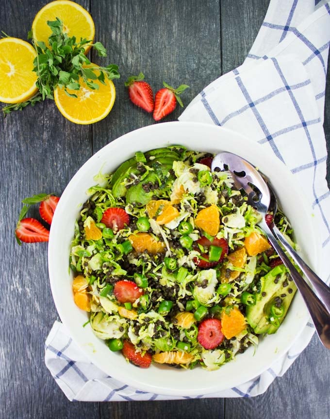 Citrus Lentil Salad with Shredded Brussel Sprouts, orange segment, strawberries and apples served in a white salad bowl with some fruit and fresh herbs in the background