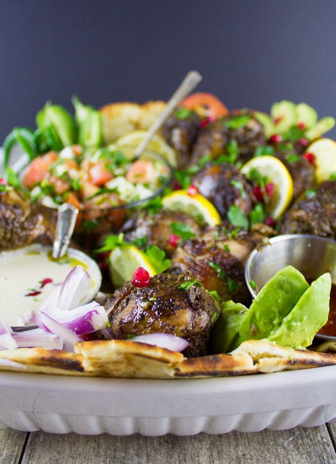 side view of Chicken Shawarma Platter served on charred naan bread with Tahini Sauce, Harissa Sauce and Tomato Feta salad in small dishes nestled in the chicken 