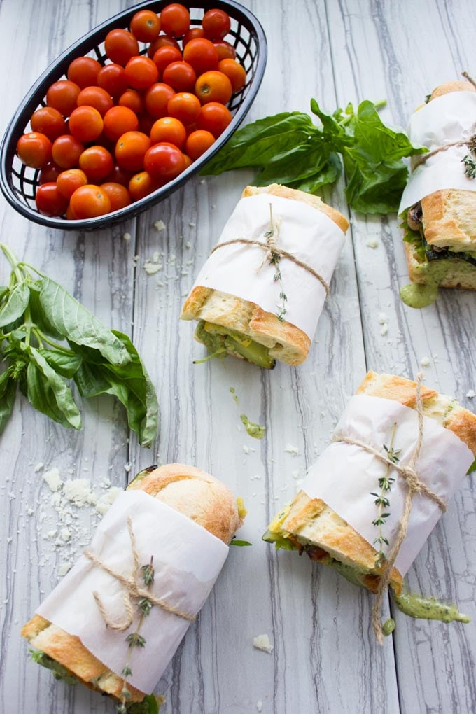 overhead shot of four Roasted Veggie Sandwiches with Basil Sauce on a white wood tabletop with cocktail tomatoes and basil on the side