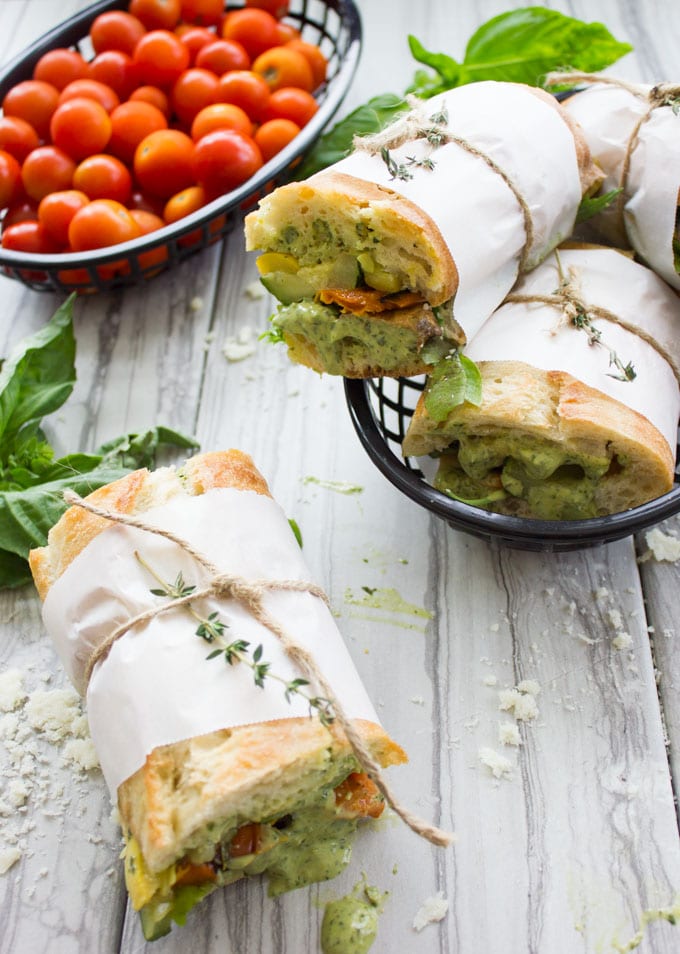 oven-roasted veggie sandwiches with basil sauce on a white rustic table with a basket of tomatoes in the background