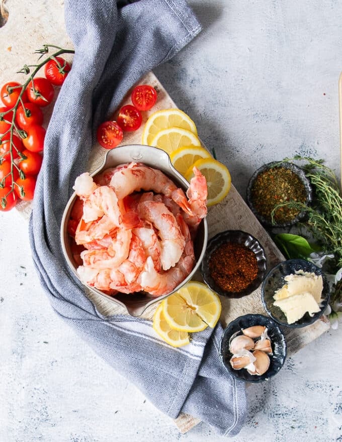 Ingredients to make shrimp pasta including raw shrimp, a bowl of seasoning,  some lemon slices, garlic slices, fresh herbs, baby tomatoes 