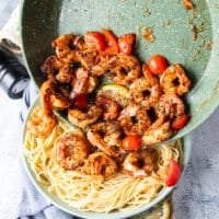 The cooked shrimp and sauce is being poured over a plate of cooked pasta to toss around