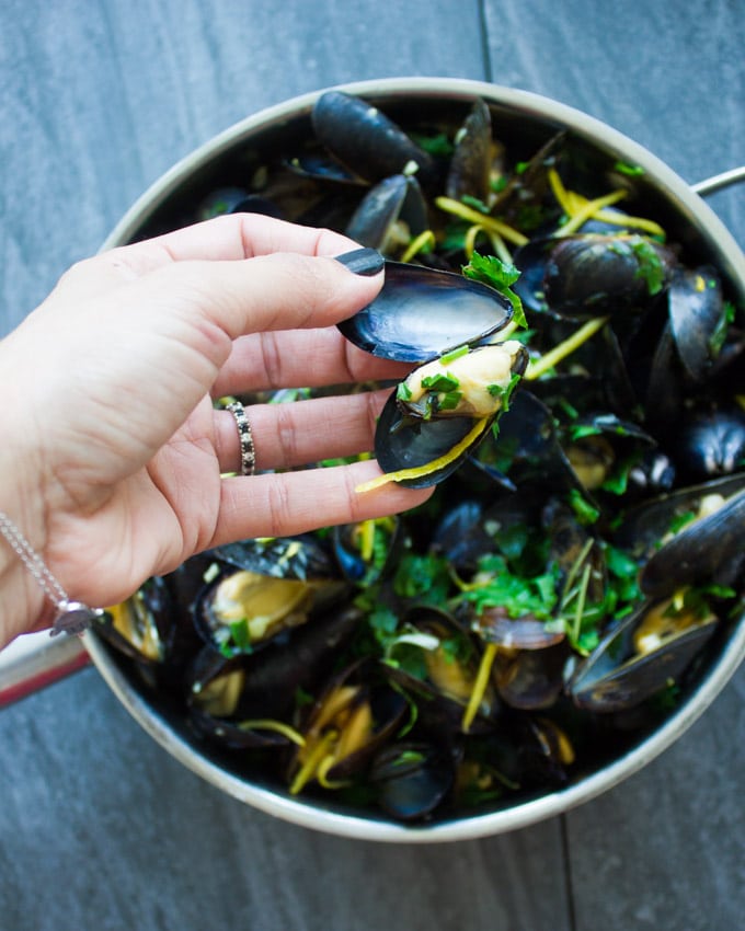 A hand showing a cooked mussel out of its' shell ready to be eaten right away over a skillet of cooked mussels.