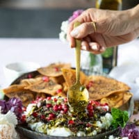 A hand spooning over the toppings over the spread out labneh over a serving plate with the pita chips