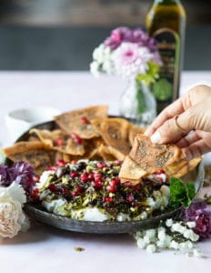 A plate of labneh dip topped with zaatar and a hand holding pita chips dipping it in the dip