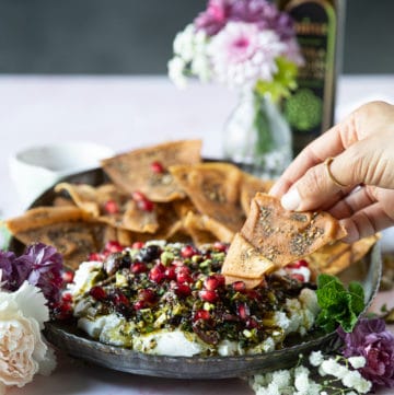 A plate of labneh dip topped with zaatar and a hand holding pita chips dipping it in the dip