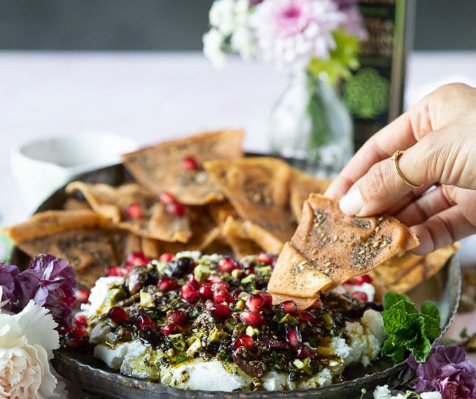 A plate of labneh dip topped with zaatar and a hand holding pita chips dipping it in the dip