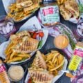 a top view table spread showing four plates with four sandwiches split in half served with some fries and cold drinks