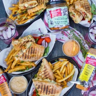 a top view table spread showing four plates with four sandwiches split in half served with some fries and cold drinks