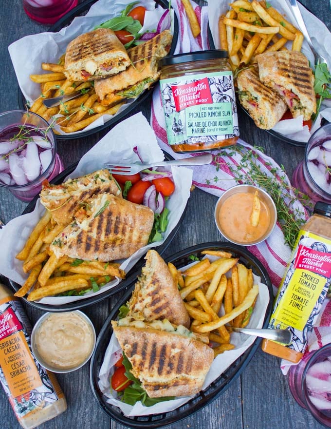 a top view table spread showing four plates with four sandwiches split in half served with some fries and cold drinks