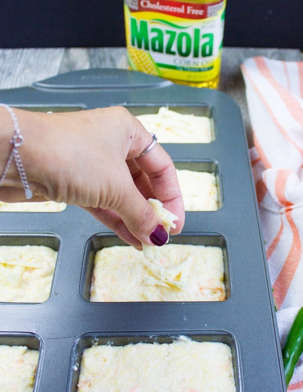 A hand sprinkling some grated cheese on the corn bread muffins batter before baking