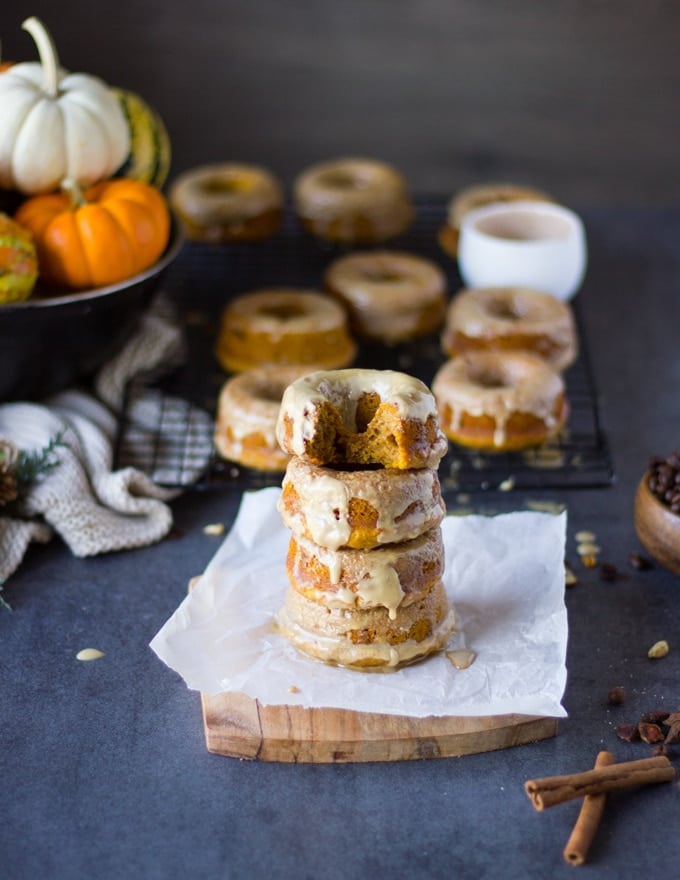 stacked up pumpkin donuts on a paper with remaining pumpkin donuts in the background and a cup of espresso