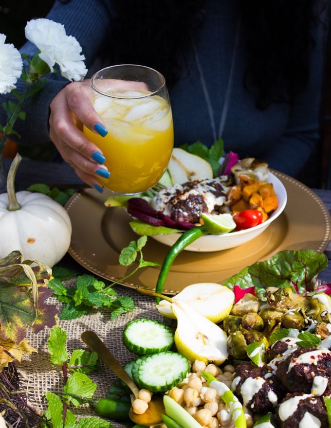 A hand holding a cup of juice over the bowl of lamb meatballs and fall roast veggies