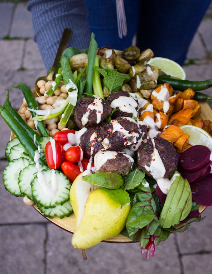 A hand holding a bowl of lamb meatballs, roast veggies, avocados, cucumbers, tomatoes and tahini sauce