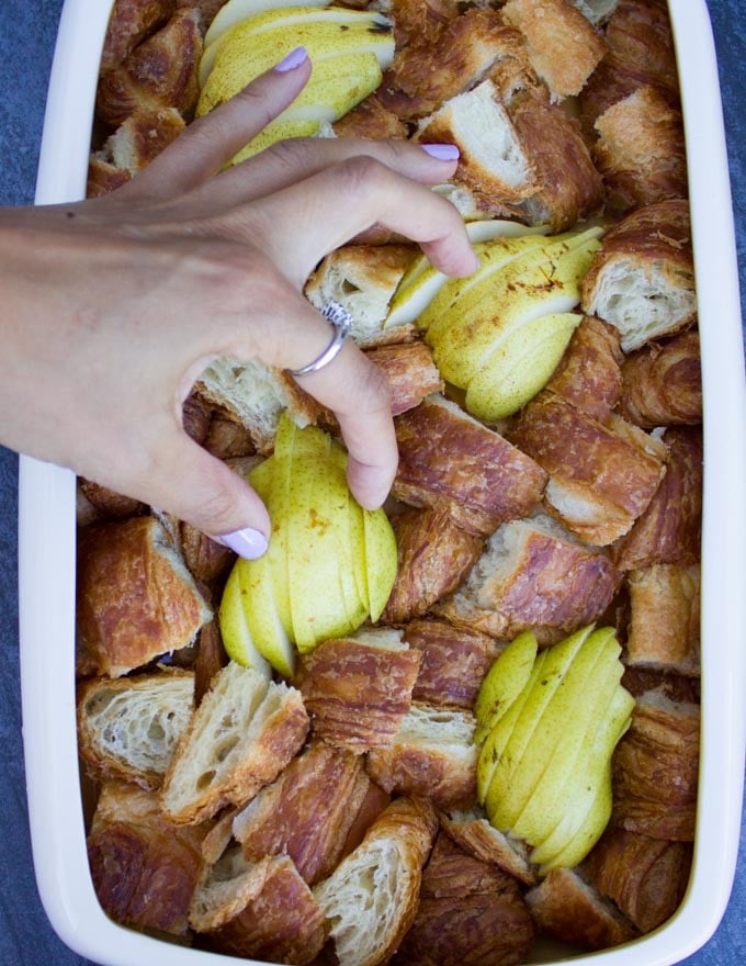 a hand placing some sliced pears in between the croissant cubes