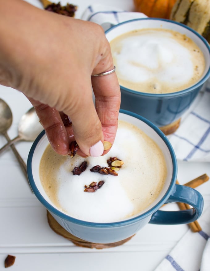 A hand sprinkling chocolate pumpkin crunch over the cup of pumpkin spice oatmeal latte