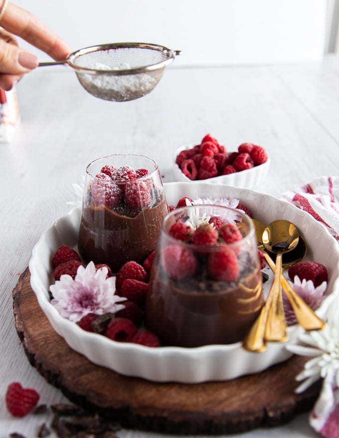 A hand dusting confectioner sugar over chocolate pudding topped with fresh raspberries