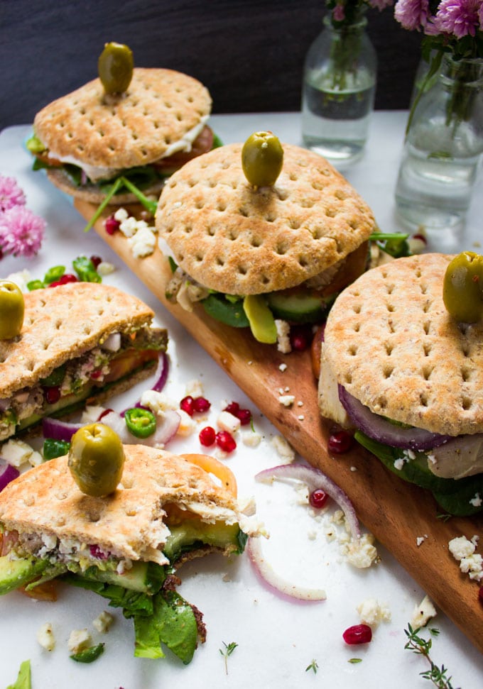 Mediterranean turkey sandwiches over a cutting board on a white granite surface with some flowers, sprinkles of feta cheese, pomegrante arils and a bitten sandwich next to them.