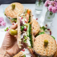 A hand holding a cut up turkey sandwich showing all the layers of turkey, cucumbers, spinach, tomatoes and feta cheese with a green olive skewered on the top.