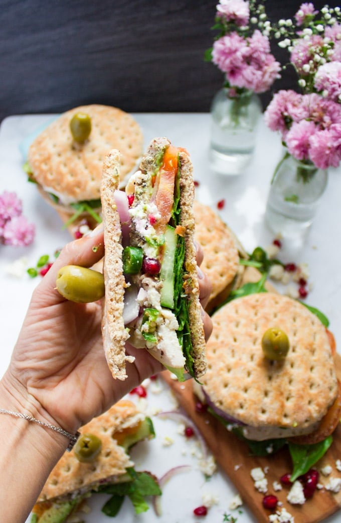 A hand holding a cut up turkey sandwich showing all the layers of turkey, cucumbers, spinach, tomatoes and feta cheese with a green olive skewered on the top.