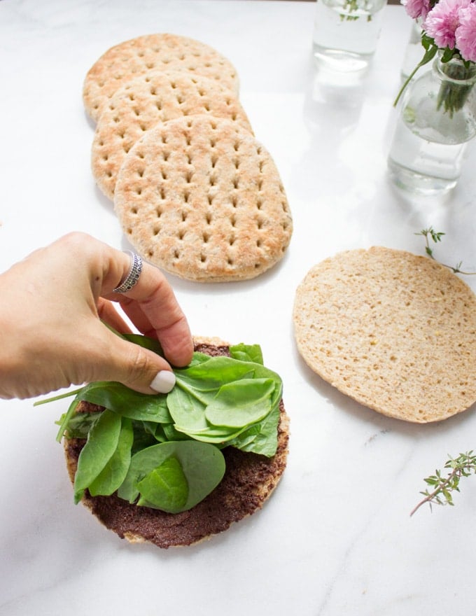 A hand piling up some fresh spinach leaves over the tapenade spread pita bread bun