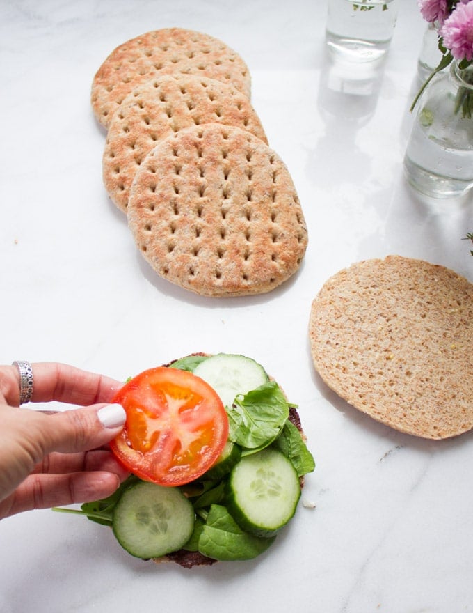A hand adding sliced tomatoes over the pita bread bun which is already layered with tapenade, spinach and cucumbers
