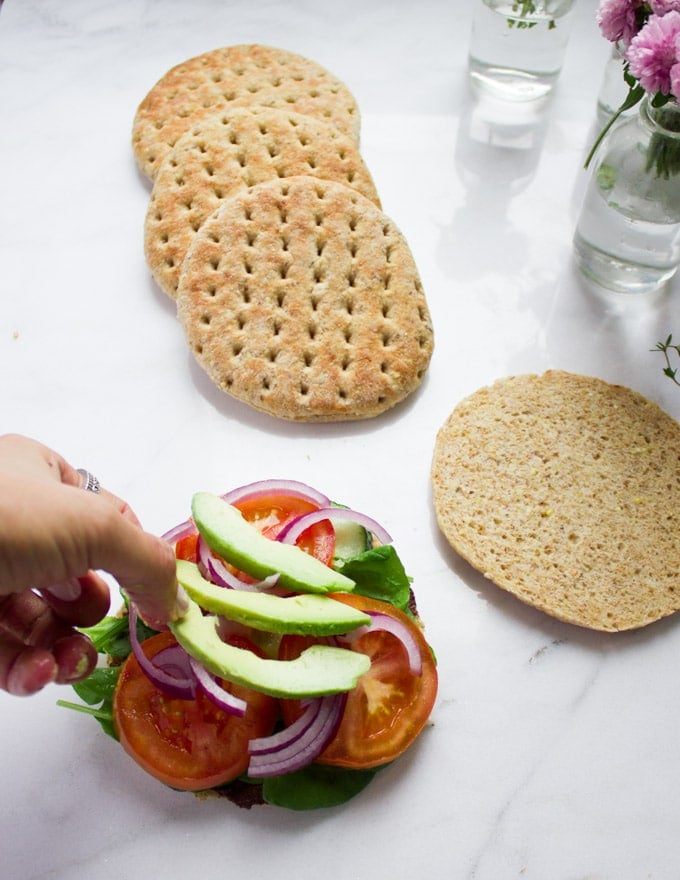 A hand adding sliced avocados and red onions on top of a pita bun with tomatoes, spinach, cucumbers and olive tapenade spread