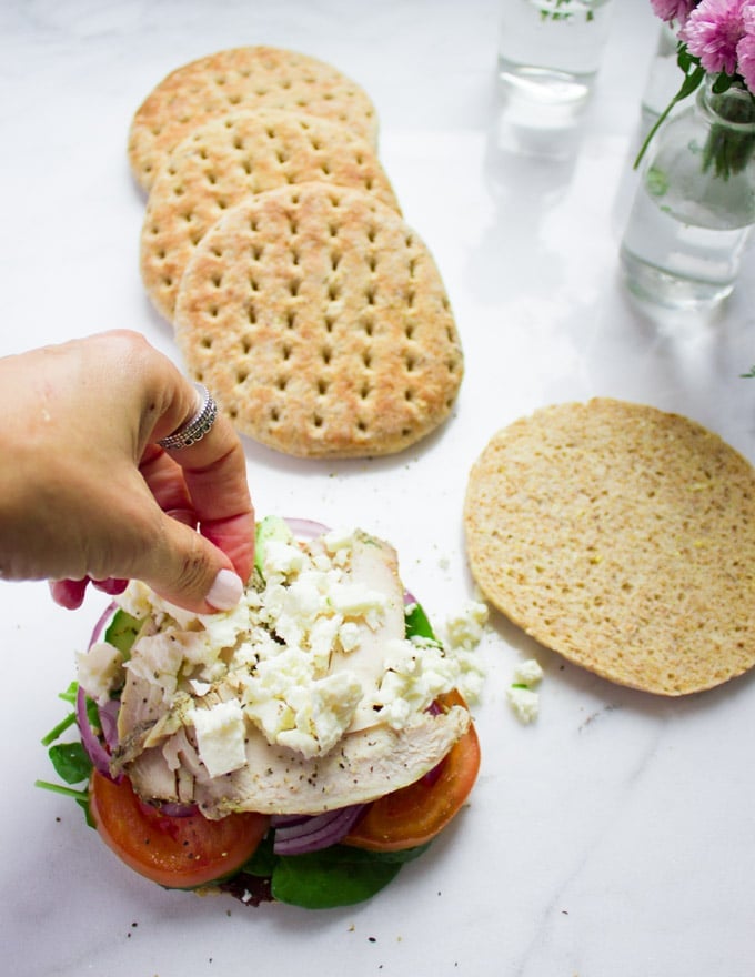 A hand spinkling feta cheese over the turkey in the sandwich over pita buns, avocados, tomatoes, red onions, cucumbers and spinach