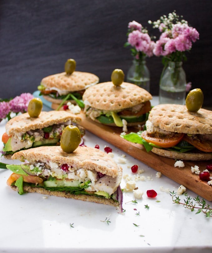 A cut up turkey sandwich on a white granite surface with the remaining turkey sandwiches behind it showing faintly. A green olive is skewered over every sandwich top. Some feta cheese is crumbled around it.