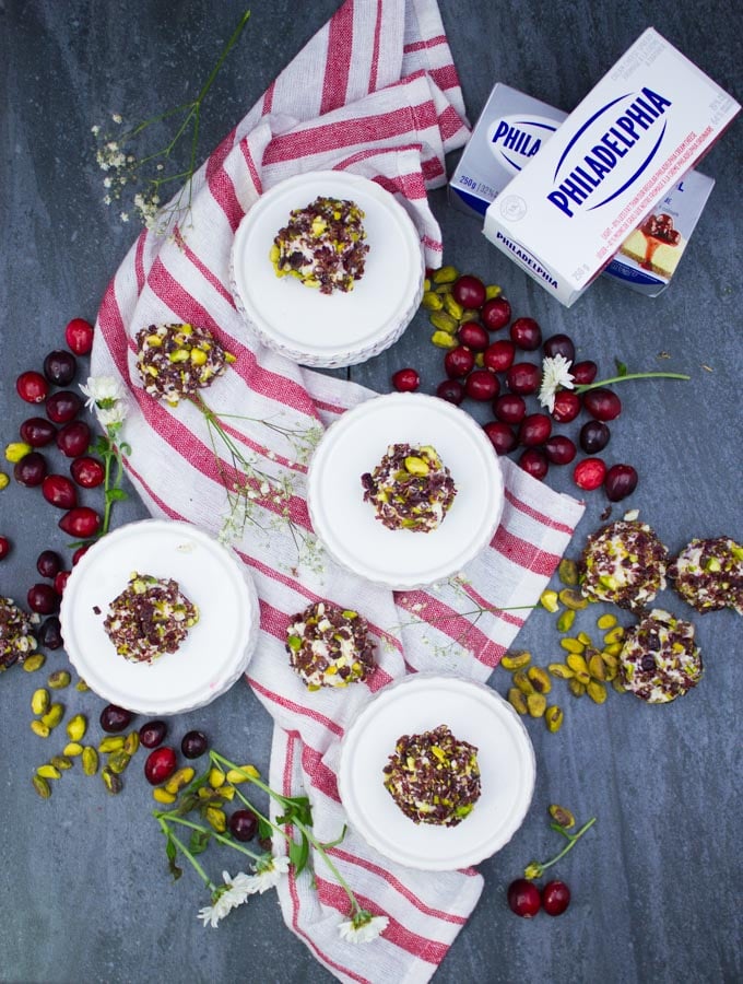 Tops view of mini cheesecake bites over small serving platters and a tea towel with more mini cheesecake bites around it. Some fresh cranberries and pistachios around it.