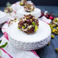 A single mini cheesecake coated in cranberries and pistachios over a small serving plate. A tea towel is around it with scattered pistachios.