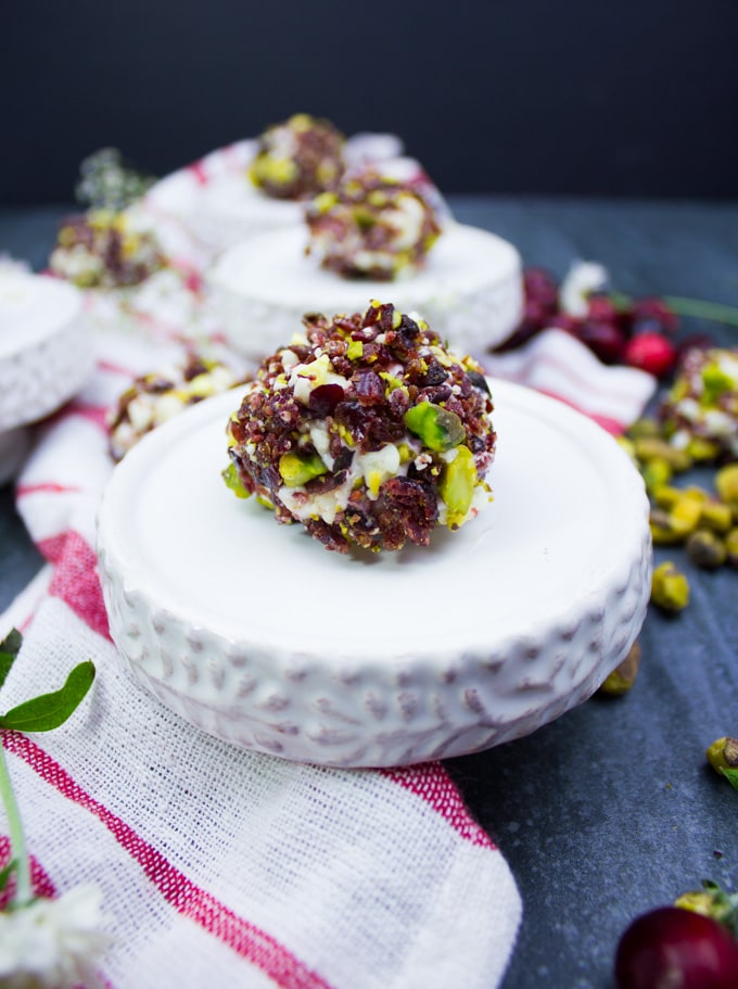 A single mini cheesecake coated in cranberries and pistachios over a small serving plate. A tea towel is around it with scattered pistachios.
