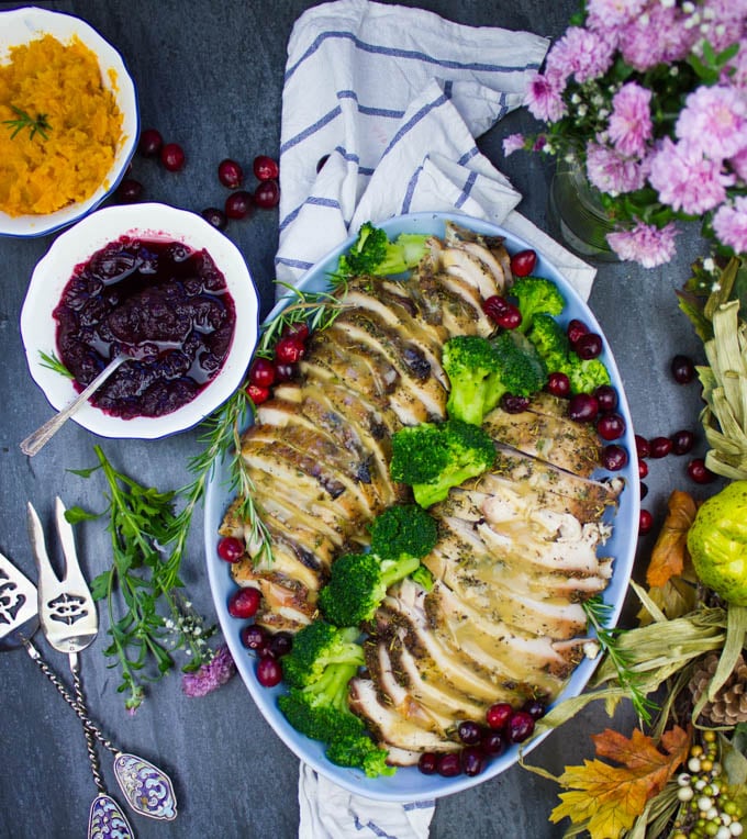 A whole set up showing the whole platter of sliced roast turkey breasts, broccoli florets, a bowl of cranberry sauce, a bowl of mashed sweet potatoes, serving spoons and some fresh cranberries for decor