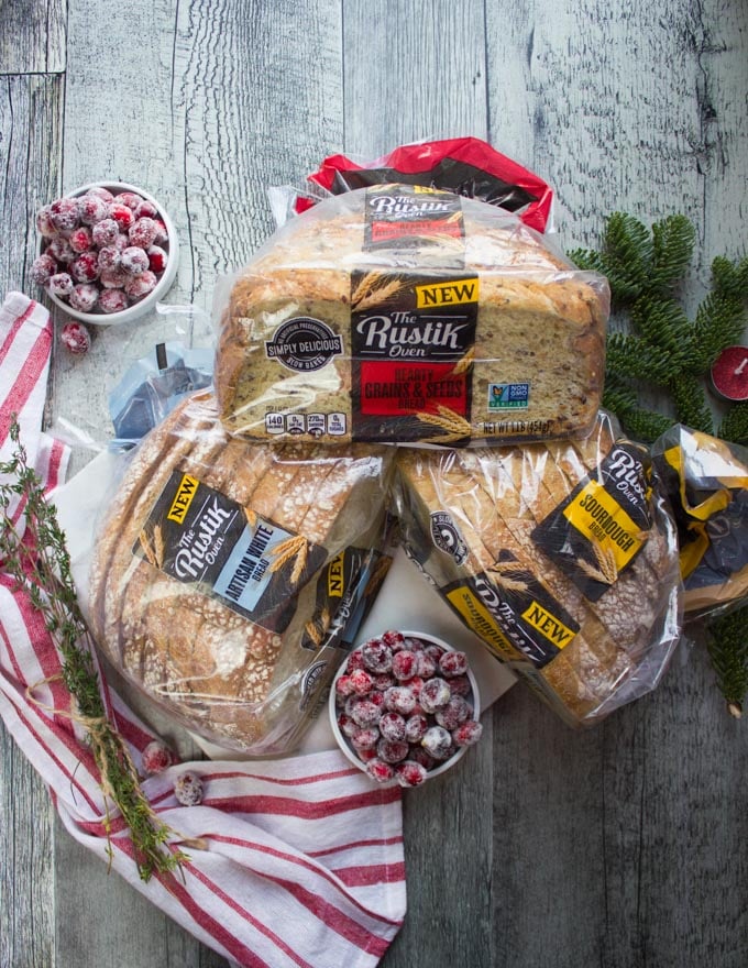 3 bags of The Rustik Oven Breads over a wooden table and a red tea towel with a bowl of frosted cranberries on the side