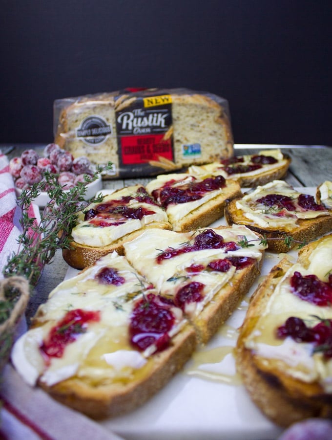 Side view of some baked brie toasts with a tea towel and a bag of the rustik oven bread in the back