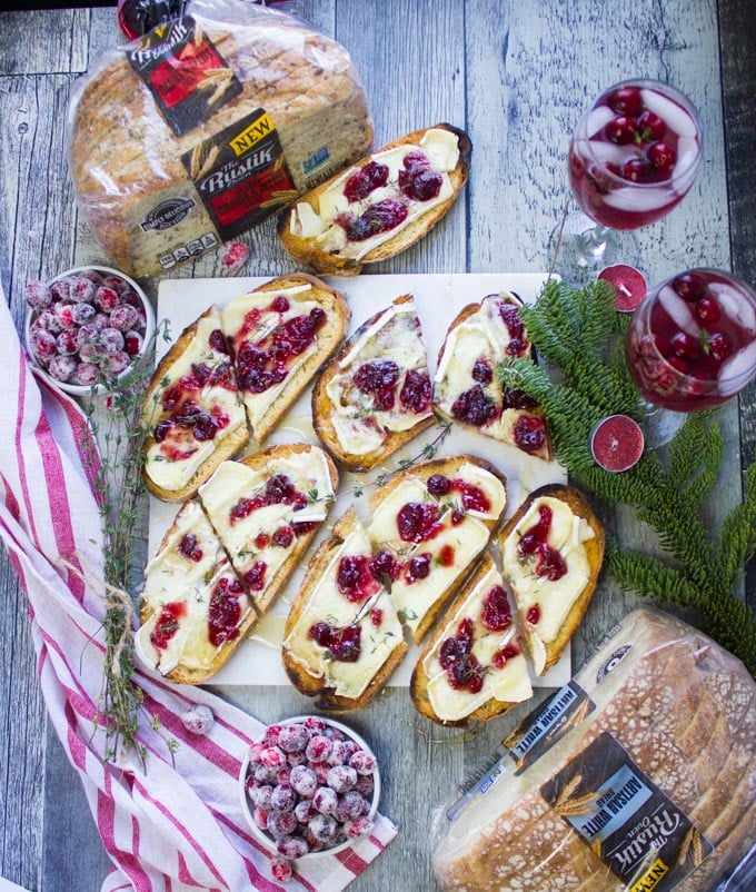 Top view of all the finished baked brie toasts on a white marble over a wooden table with bags of bread around it, some cranberry cocktails and frosted cranberries