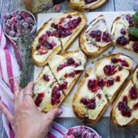 A hand picking up a half toast of baked brie from the mable of all the baked brie toats surrounded by a white and red tea towel