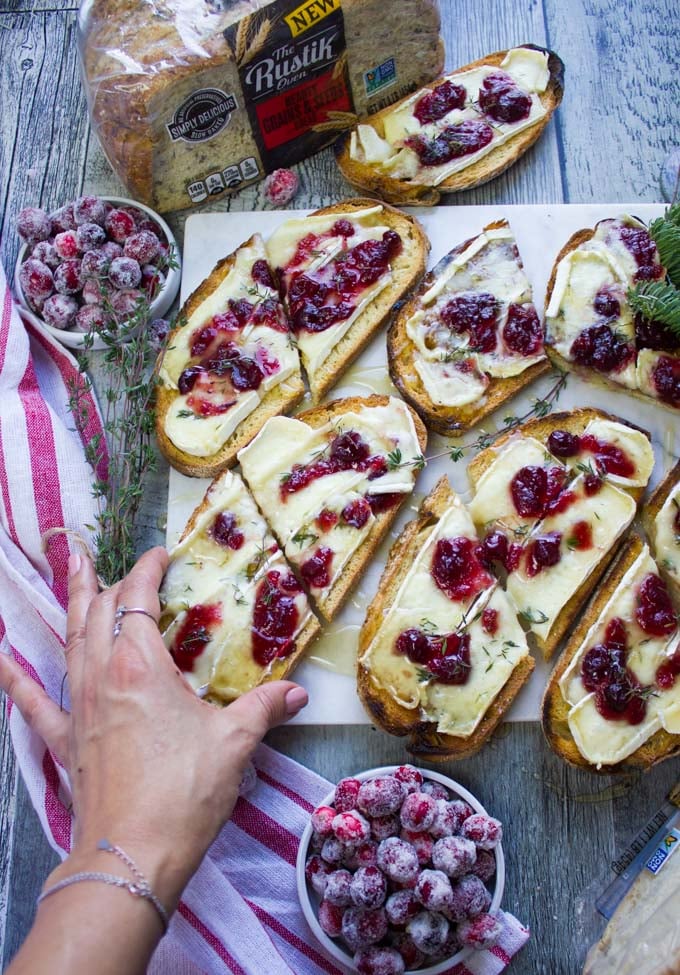 A hand picking up a half toast of baked brie from the mable of all the baked brie toats surrounded by a white and red tea towel