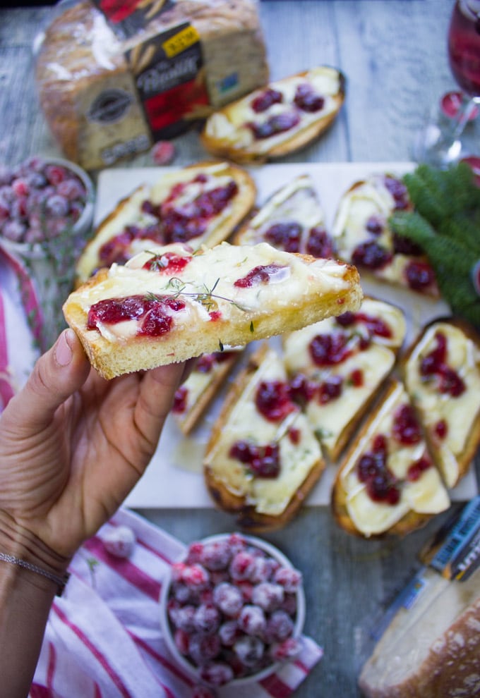 A hand holding hald a baked brie toast up close showing the texture of the bread and the layering of brie cheese, cranberry sauce and honey