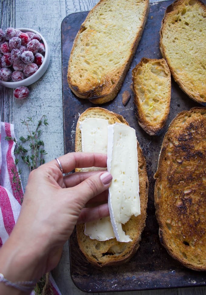 A hand placing slices of brie cheese over toasted bread on a metal sheet pan