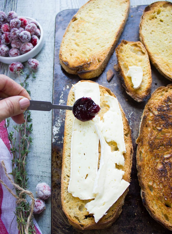 A spooning assembling the baked brie toast by adding some cranberry sauce over the brie before baking