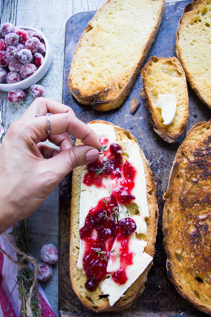 A hand topping the brie and cranberry sauce over toast with some fresh thyme leaves