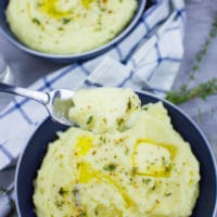 A fork holding some creamy mashed potatoes over a big blue plate of mashed potatoes and a white towel.