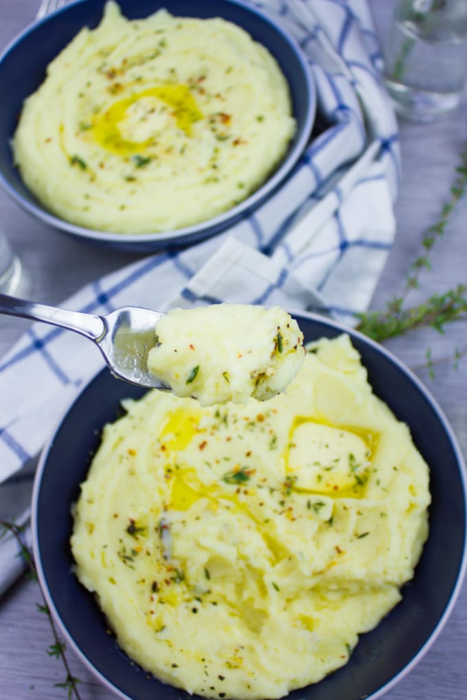 A fork holding some creamy mashed potatoes over a big blue plate of mashed potatoes and a white towel.