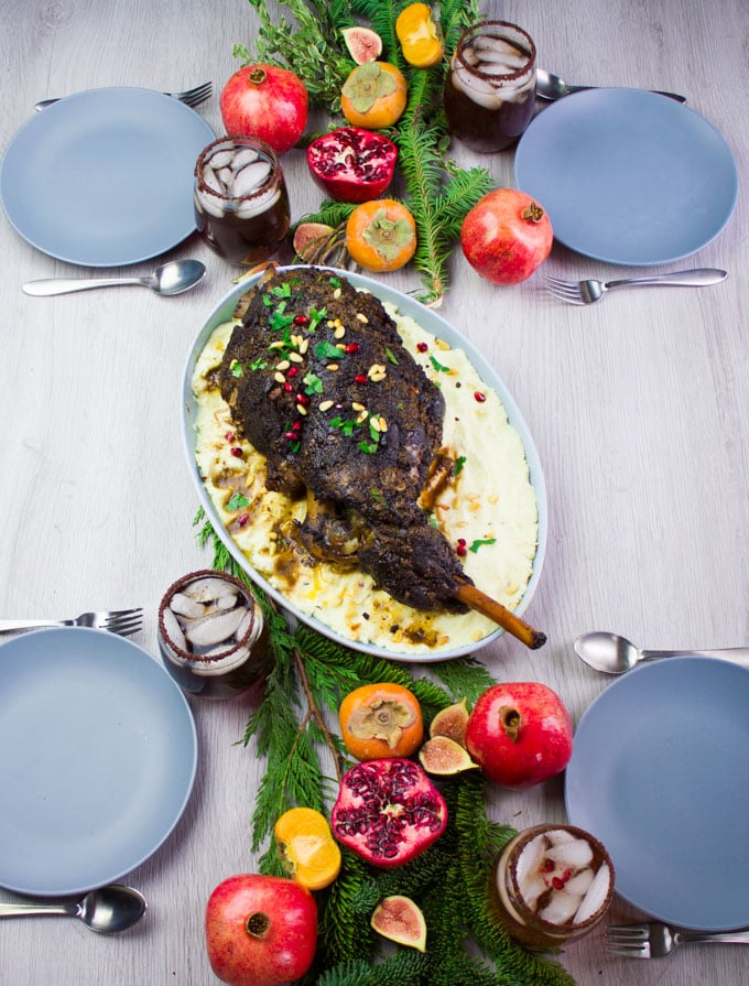 A table with four plates and drinks and a main dish of leg of lamb with mashed potatoes in the middle surrounded by pomegranates and greens