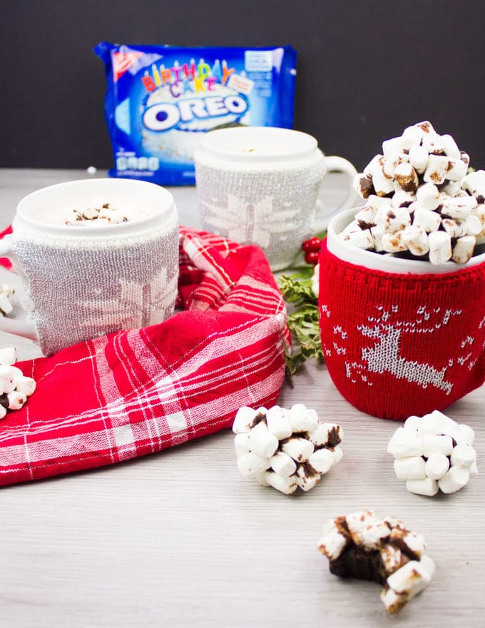 A side view of the mug holding several OREO cookie balls, and an OREO pack at the background and two hot chocolate cups at the back.