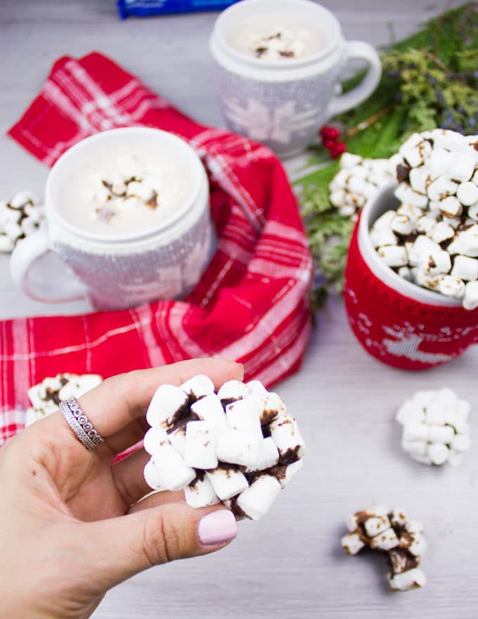 A hand Holding an OREO cookie balls coated in marshmallows up close, with two hot chocolate drinks at the back.