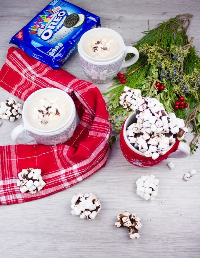 A spread of a red towel, a holiday wreath and two hot chocolate cups, topped with OREO cookie balls, an OREO pack at the background and a cup filled with OREO cookie balls