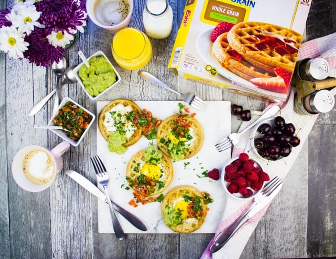 Top view of a breakfast table with egg in a hole wafles topped with guacamole and salsa, a box of waffles, some coffee, orange juice and berries to eat.